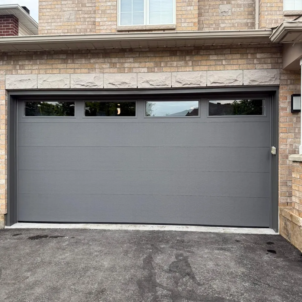 Grey flush-panel double garage door with four narrow top windows in a brick home.